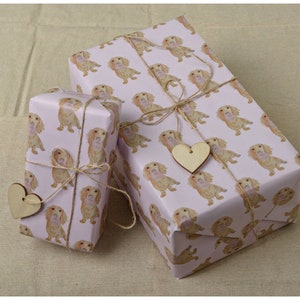 May include: Two wrapped gifts with light pink paper featuring a repeating pattern of golden Cocker Spaniel illustrations. The gifts are tied with twine and adorned with wooden heart-shaped tags. The gifts are on a beige surface.