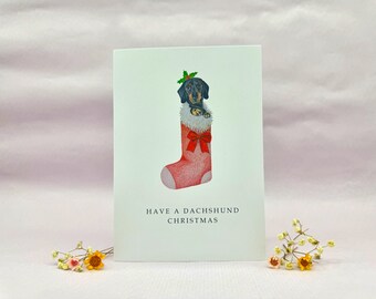 May include: A Christmas card featuring a dachshund dog in a red stocking with a white cuff and a red bow. The dog is wearing a holly wreath. The text "HAVE A DACHSHUND CHRISTMAS" is printed below the illustration.
