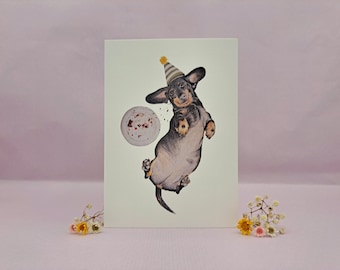 May include: A cream-coloured greeting card with a watercolour illustration of a dachshund wearing a party hat. The dog is mid-air, with a cake nearby. The image has a celebratory theme.