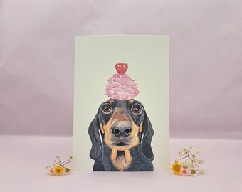 May include: A greeting card showing a dachshund with a pink cupcake on its head, topped with a red cherry. The dog has black and brown fur and is set against a light green backdrop.