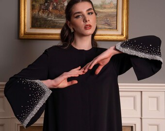 Könnte beinhalten: Eine schwarze Abaya mit langen, weit ausgestellten Ärmeln, die an den Manschetten mit aufwendigen silbernen Kristallverzierungen versehen sind. Das Kleidungsstück hat einen schlichten Rundhalsausschnitt und besteht durchgehend aus schwarzem Stoff.