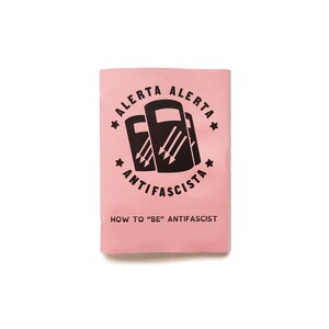 May include: A pink pamphlet with the text "Alerta Alerta Antifascista" and a black illustration of three shields with arrows. The text "How to "Be" Antifascist" is printed below the illustration.