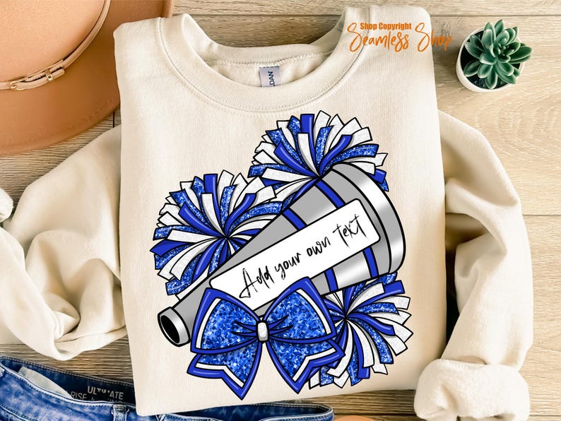 K&ouml;nnte beinhalten: Cremefarbenes Sweatshirt mit einem Cheerleading-Design. Die Grafik zeigt ein silbernes Megaphon, blau-wei&szlig;e Pompons und eine blaue Schleife. Der Text "Add your own text" wird auf einem wei&szlig;en Rechteck angezeigt.