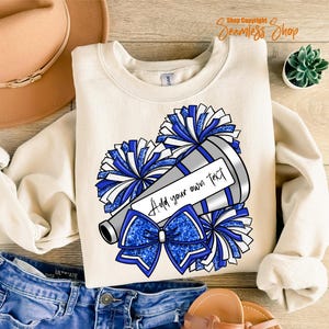 K&ouml;nnte beinhalten: Cremefarbenes Sweatshirt mit einer Grafik eines Megaphons, Pompons und einer Schleife in Blau- und Silbert&ouml;nen. Das Megaphon hat einen Bereich, in dem Sie Ihren eigenen Text hinzuf&uuml;gen k&ouml;nnen. Das Sweatshirt wird mit Jeans und einem beigen Hut kombiniert.
