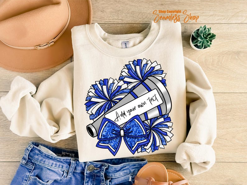 K&ouml;nnte beinhalten: Cremefarbenes Sweatshirt mit einer Grafik eines Megaphons, Pompons und einer Schleife in Blau- und Silbert&ouml;nen. Das Megaphon hat einen Bereich, in dem Sie Ihren eigenen Text hinzuf&uuml;gen k&ouml;nnen. Das Sweatshirt wird mit Jeans und einem beigen Hut kombiniert.