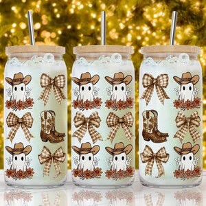 Op de afbeelding: Drie glazen bekers met houten deksels en metalen rietjes. Elke beker heeft een herhalend patroon van een cartoongeest met een cowboyhoed, cowboylaarzen en een geruite strik. De achtergrond is een bokeh van gouden lichten.