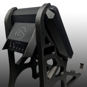 May include: A black electronic device with a geometric design, mounted on a black stand. The device has a rectangular body and a series of ports. The stand has an open, architectural structure.