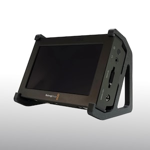 May include: A black video monitor with a black frame. The monitor has a black screen and is angled slightly upward. The frame has a built-in stand and several ports and buttons on the side. The text "Blackmagic Design" is visible.