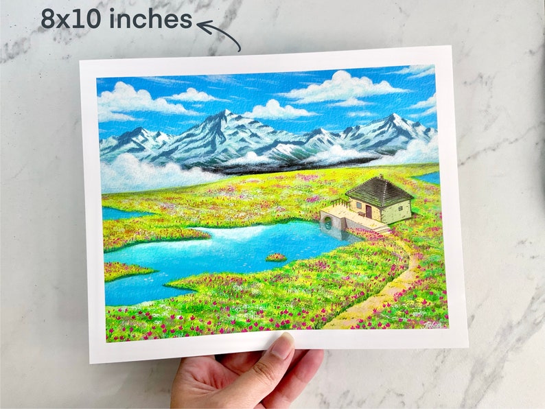 May include: An 8x10 inch print of a watercolour painting depicting a small cottage with a water wheel in a field of flowers. The cottage is surrounded by a blue lake and snow-capped mountains in the background.