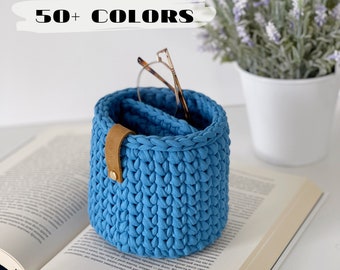 Puede incluir: Una cesta de almacenamiento de crochet azul con una correa de cuero marrón y un botón de metal. La cesta está sentada en un libro y tiene gafas dentro.