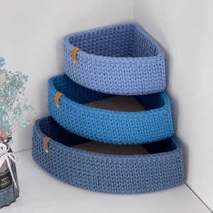 May include: Three blue crocheted corner shelves with brown leather handles. The shelves are stacked on top of each other.