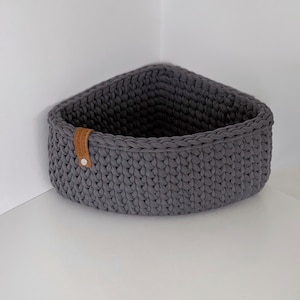 May include: A grey crocheted corner basket with a brown leather label. The basket is made of thick yarn and has a textured surface.