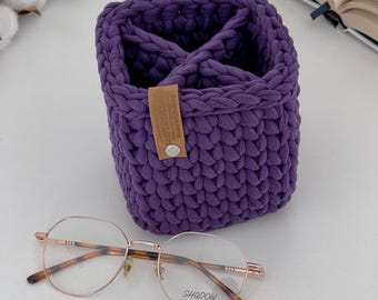 Puede incluir: Cesta de almacenamiento de color morado hecha a crochet con una etiqueta de cuero marrón que dice "Handmade with Love". Un par de gafas de montura dorada con lentes transparentes está en primer plano.