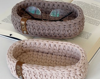 May include: Two crocheted oval-shaped baskets with leather labels that read "JOLIE". The top basket is a light brown color and the bottom basket is a light beige color.