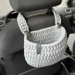 Op de afbeelding: Grijze gehaakte auto-organizer met een leren label met de tekst "CRAFT BY ANNE", hangend aan de hoofdsteun van een autostoel.