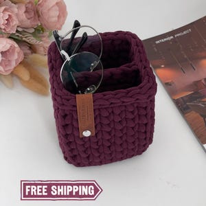 Puede incluir: Una cesta de ganchillo burdeos con dos compartimentos, que contiene un par de gafas. La cesta tiene una etiqueta de cuero que dice "RAFTBYANNE". La imagen también incluye una revista y flores rosas.