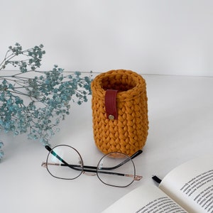 May include: A crocheted mustard yellow pencil holder with a red leather strap and a silver button. The holder is sitting on a white surface next to a pair of eyeglasses and a book.