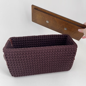 Puede incluir: Una cesta rectangular de ganchillo morado oscuro con una tapa de madera a juego. La cesta tiene un aspecto texturizado y tejido y asas de cuero. La tapa es de madera oscura con dos cierres magnéticos plateados. La cesta está diseñada para el almacenamiento.