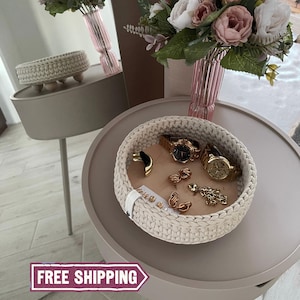 May include: A cream-colored, crocheted basket filled with gold watches and jewelry. The round basket sits on a light brown table, with a smaller basket on a side table in the background. A vase of flowers is also visible.