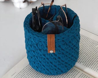 Puede incluir: Un contenedor cilíndrico de crochet azul turquesa con una etiqueta de cuero marrón que dice "CRAFTBYANNED made with love" y cuatro pares de gafas en su interior.