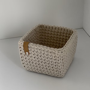 May include: A square, cream-coloured crochet basket with a braided top edge. A small, brown leather tag is attached to the side with a silver button. The basket is made of thick, soft yarn and is empty.