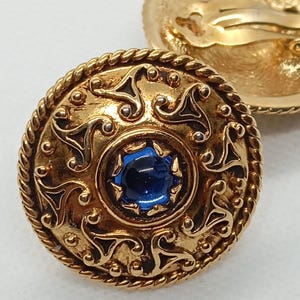 Pode incluir: Um par de brincos de clip dourados com uma pedra azul no centro de cada brinco. Os brincos têm um design detalhado com uma borda de corda e padrões intrincados.