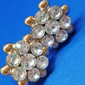 Puede incluir: Un par de pendientes de tono dorado con diamantes de imitación transparentes. Cada pendiente tiene un grupo de siete diamantes de imitación dispuestos en un patrón circular.