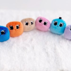 May include: Six plush toys shaped like cubes with different colours and facial expressions. The colours are blue, orange, brown, pink, light blue, and purple.