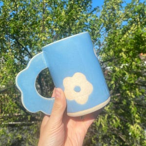 Könnte beinhalten: Hellblaue Keramiktasse mit blütenförmigem Griff und cremefarbenem Blumendesign. Die Tasse hat einen abgerundeten Boden und wird vor dem Hintergrund von grünem Laub und blauem Himmel gehalten.