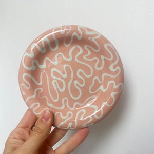 May include: A small, round, ceramic dish with a light pink base and a white, abstract, squiggly pattern. The dish is held by a hand against a white background. The dish is likely 10-15 cm in diameter.
