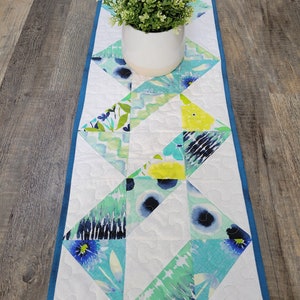 Peut inclure: Un chemin de table blanc avec une bordure bleue et un motif géométrique de fleurs aquatiques, vertes, jaunes et noires.