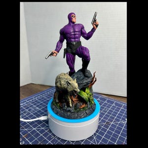 May include: A purple figurine of a muscular figure holding two pistols, standing on a rock formation. A wolf-like creature is at the base, surrounded by foliage. The figurine is on a white and blue rotating display stand.