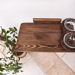 May include: A dark brown wooden pet food station with two glass bowls attached to the side. The station is mounted to a white wall.