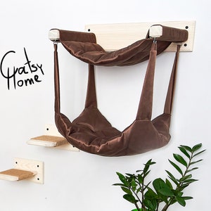 May include: A brown cat hammock with a wooden platform and two smaller platforms. The hammock is suspended from the platform by two ropes. The platform is mounted to a white wall. The text "Gratsy Home" is written on the wall.