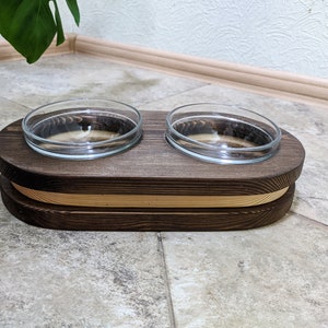 May include: A wooden pet food bowl stand with two glass bowls. The stand is dark brown with a light brown accent band. The bowls are clear glass.