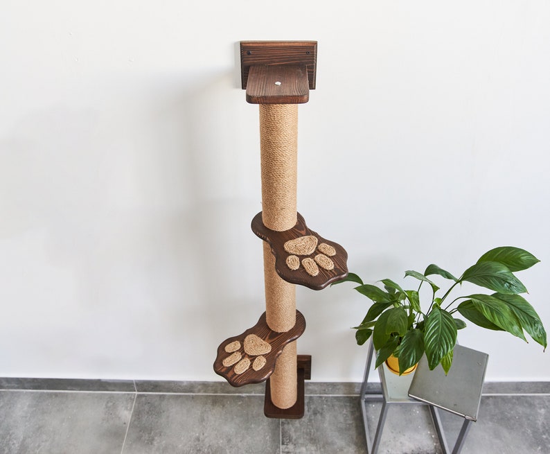 May include: A brown wooden cat scratching post with three platforms. The platforms are shaped like paw prints and are covered in sisal rope. The post is mounted to a wall.