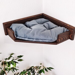 May include: A dark brown wooden corner shelf with a gray cushion. The shelf is mounted on a white wall.