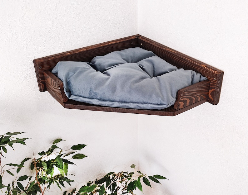 May include: A dark brown wooden corner shelf with a gray cushion. The shelf is mounted on a white wall.