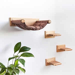 Pode incluir: Um conjunto de tr&ecirc;s prateleiras de madeira para gatos, incluindo uma cama tipo rede. As prateleiras s&atilde;o feitas de madeira natural e t&ecirc;m um acabamento castanho claro. A cama rede &eacute; castanha e tem um interior macio e acolchoado. As prateleiras s&atilde;o montadas numa parede branca.