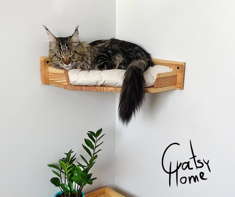 May include: A wooden cat bed mounted in a corner of a room. The bed is made of light wood and has a white cushion. A brown tabby cat is sleeping on the bed. The text "Gratsy Home" is written in black on the wall.