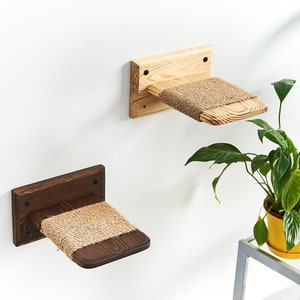 May include: Two wooden cat shelves mounted on a white wall. One shelf is light-colored wood, the other is dark brown. Both have a woven sisal mat for the cat to rest on. The shelves are designed to be mounted on a wall.