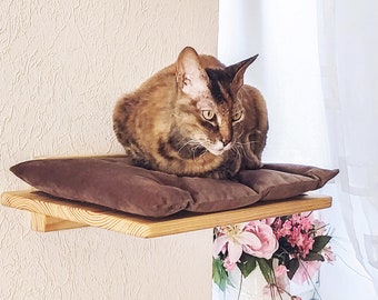 May include: A brown and black cat sits on a brown cushion on a wooden shelf. The shelf is mounted to a white wall.