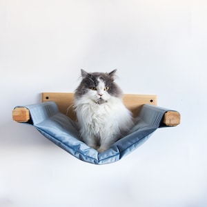 May include: A gray and white cat sits in a blue hammock bed attached to a wooden wall mount. The hammock is made of soft, plush fabric and is designed for cats to relax and sleep in.
