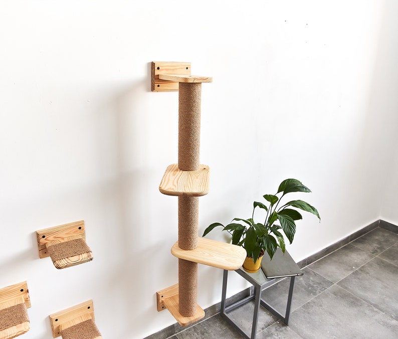 May include: A wooden cat tree with three platforms and a scratching post. The cat tree is mounted to a white wall. The platforms are made of natural wood and the scratching post is covered in sisal rope.