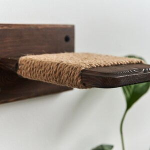 May include: A wooden cat shelf with a sisal rope surface. The shelf is mounted to a white wall and is angled slightly downward.
