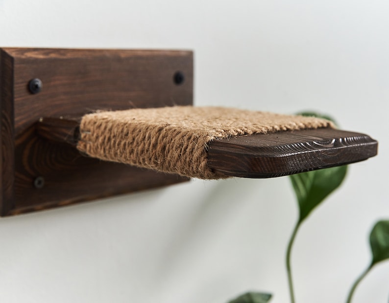 May include: A wooden cat shelf with a sisal rope surface. The shelf is mounted to a white wall and is angled slightly downward.