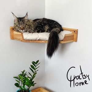 May include: A wooden cat bed mounted in a corner of a room. The bed is made of light wood and has a white cushion. A brown tabby cat is sleeping on the bed. The text "Gratsy Home" is written in black on the wall.