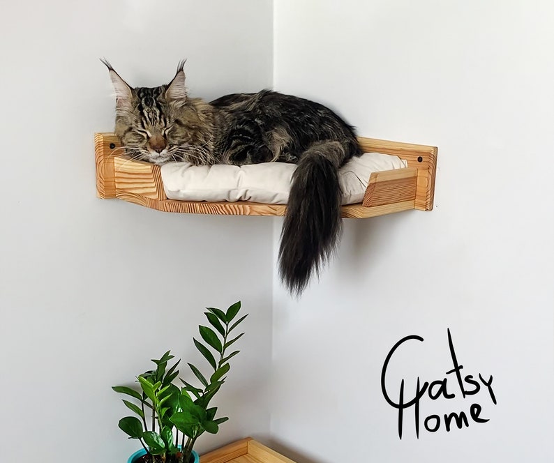 May include: A wooden cat bed mounted in a corner of a room. The bed is made of light wood and has a white cushion. A brown tabby cat is sleeping on the bed. The text "Gratsy Home" is written in black on the wall.