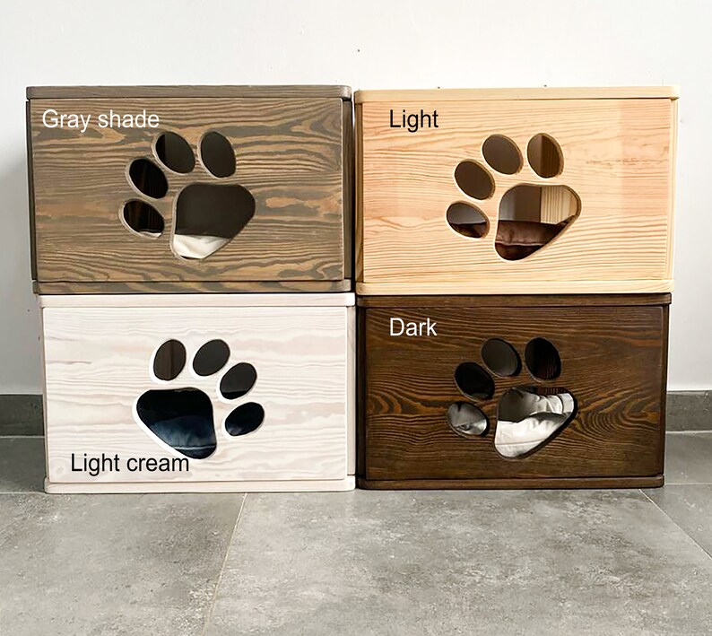 May include: Four wooden cat beds in different shades of brown, gray, and cream. Each bed has a paw-shaped opening and a soft cushion inside. The beds are labeled "Gray shade", "Light", "Light cream", and "Dark".