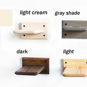 May include: Four wooden cat shelves in different colors: light cream, gray shade, dark brown, and light wood. Each shelf is mounted on a wall and has a small platform for cats to perch on.